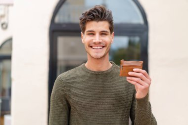 Açık havada cüzdan tutan genç, beyaz bir adam çok fazla gülümsüyor.