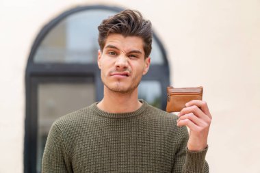 Kafkasyalı genç bir adam dışarıda üzgün bir ifadeyle bir cüzdan tutuyor.