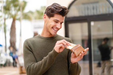 Açık havada genç beyaz bir adam elinde cüzdanla mutlu bir ifadeyle.