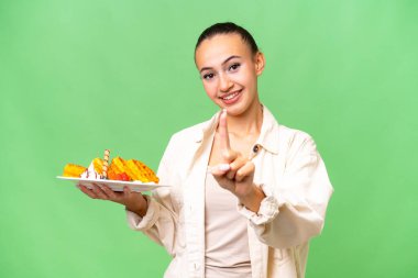 İzole edilmiş arka planda waffle tutan genç bir Arap kadın parmağını kaldırıyor ve gösteriyor.