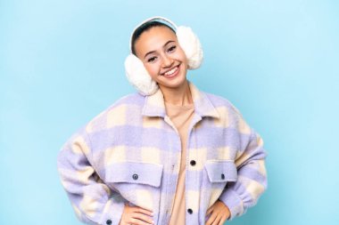 Mavi arka planda izole edilmiş, kalçasında kollarıyla poz veren ve gülümseyen genç bir Arap kadın.