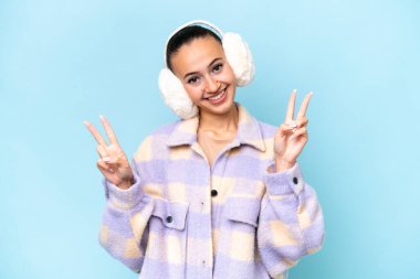 Mavi arka planda izole edilmiş kışlık manşet giyen genç bir Arap kadın zafer işaretini iki eliyle de gösteriyor.