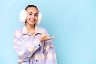 Mavi arka planda izole edilmiş kışlık manşet giyen genç bir Arap kadın gülümseyerek bir fikir sunuyor.