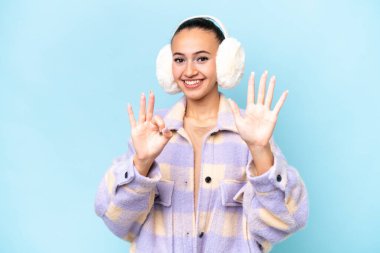 Mavi arka planda izole edilmiş kışlık kuklalar giyen genç Arap kadın parmaklarıyla sekiz tane sayıyor.