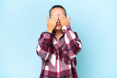 Mavi arka planda izole edilmiş genç bir Arap kadın gözlerini elleriyle kapatıyor.