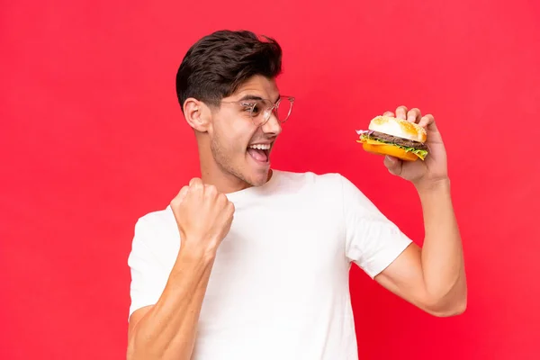 Młody Biały Mężczyzna Trzyma Hamburgera Czerwonym Tle Świętując Zwycięstwo — Zdjęcie stockowe
