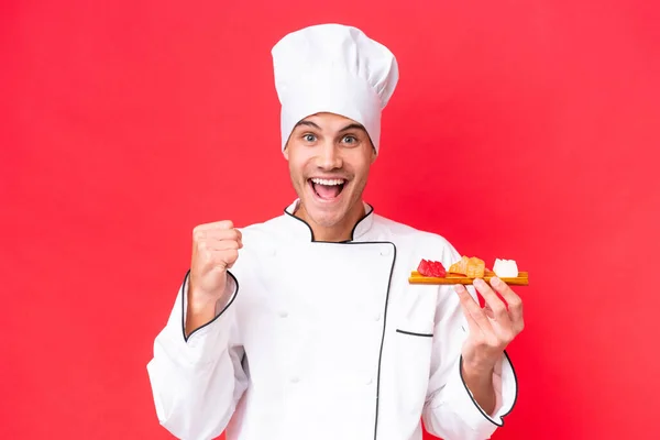 赤い背景に孤立した刺身を持つ若い白人料理人が優勝を祝う — ストック写真