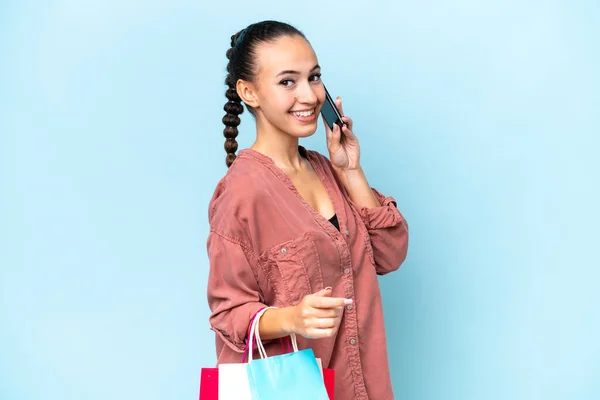 青の背景に隔離された若いアラブ人女性ショッピングバッグを保持し 彼女の携帯電話で友人を呼び出します — ストック写真