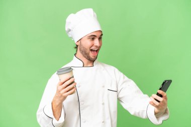 Genç, yakışıklı aşçı, izole edilmiş arka planda kahve ve bir cep telefonu tutuyor.