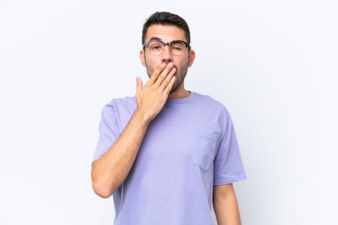 Beyaz arka planda izole edilmiş genç beyaz bir adam esniyor ve geniş bir ağzı eliyle kapatıyor.