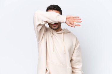 Beyaz arka planda izole edilmiş beyaz tenli genç adam gözlerini elleriyle kapatıyor.