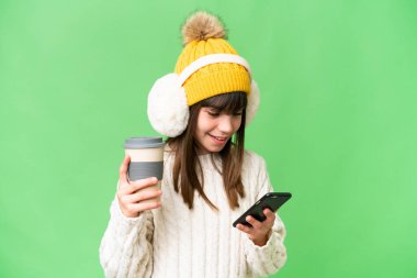 Kafkasyalı küçük bir kız izole edilmiş arka planda kışlık kuklalar takıyor. Elinde kahve ve cep telefonu var.