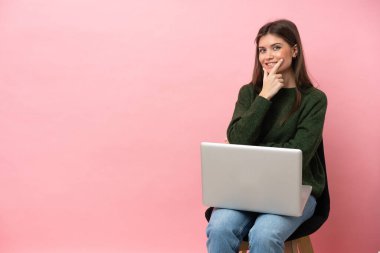 Genç beyaz kadın dizüstü bilgisayarı ile bir sandalyede oturuyor pembe arka planda izole edilmiş gülümsüyor.