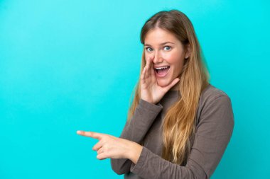 Mavi arka planda izole edilmiş genç sarışın bir kadın bir ürün sunmak ve bir şeyler fısıldamak için kenarı işaret ediyor.