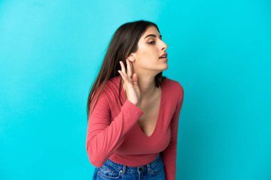 Mavi arka planda izole edilmiş genç beyaz kadın elini kulağına koyarak bir şeyler dinliyor.