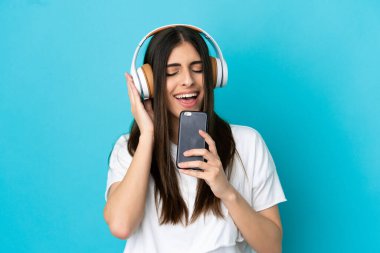 Genç beyaz kadın mavi arka planda izole edilmiş. Cep telefonuyla müzik dinliyor ve şarkı söylüyor.
