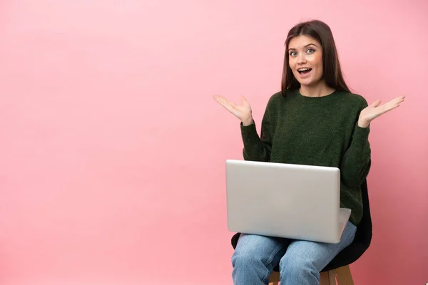 年轻的高加索女人坐在椅子上 笔记本电脑被粉色背景隔离 满脸震惊的表情 — 图库照片