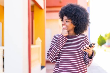 Açık havada cep telefonu kullanan Afrikalı Amerikalı bir kız bir fikir üretiyor ve ona bakıyor.