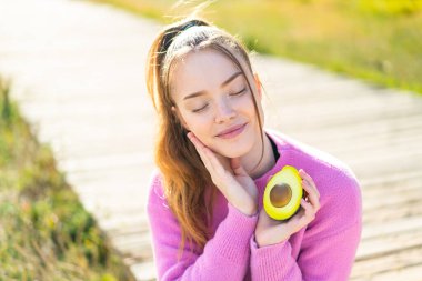 Açık havada elinde mutlu bir ifadeyle avokado tutan genç ve güzel bir kız.