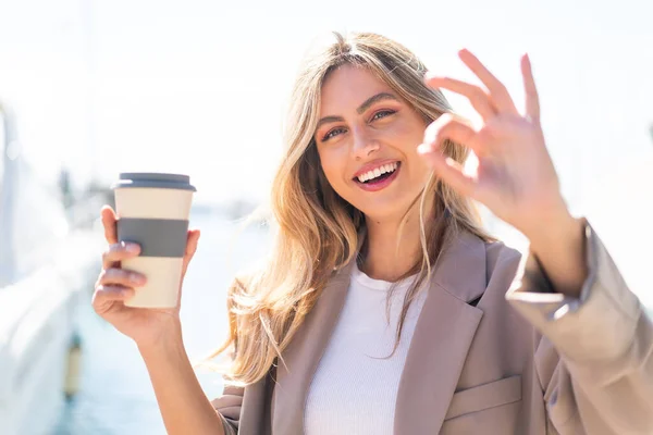 漂亮的金发碧眼的乌拉圭女人在户外端着一杯带咖啡 用手指示意好了 — 图库照片