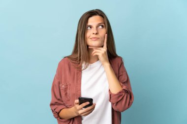 Genç Slovak kadın mavi arka planda cep telefonu kullanıyor ve düşünüyor.