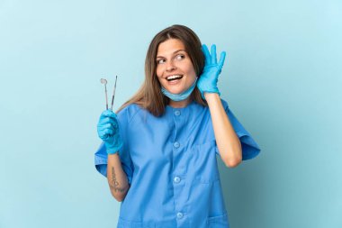 Slovak dişçi, mavi arka planda izole araçlar tutuyor. Kulağın üzerine elini koyarak bir şey dinliyor.