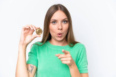 Beyaz arka planda izole edilmiş Bitcoin tutan güzel sarışın kadın şaşırmış ve ön tarafı işaret ediyor.