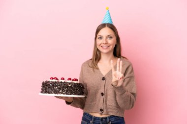 Elinde doğum günü pastası tutan genç Litvanyalı kadın pembe arka planda izole edilmiş mutlu ve parmaklarıyla üçe kadar sayıyor.