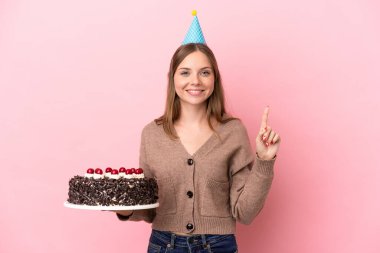 Elinde doğum günü pastası tutan Litvanyalı genç bir kadın pembe arka planda izole edilmiş ve en iyinin işareti olarak parmağını kaldırıyor.