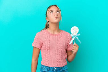 Küçük beyaz kız elinde lolipopla mavi arka planda izole edilmiş bir şekilde yukarı bakıyor.