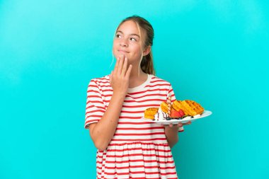 Mavi arka planda gözleme tutan küçük beyaz kız gülümserken yukarı bakıyor.
