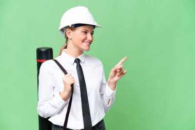 Young architect caucasian woman with helmet and holding blueprints over isolated background pointing finger to the side and presenting a product
