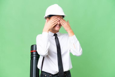 Young architect caucasian woman with helmet and holding blueprints over isolated background covering eyes by hands