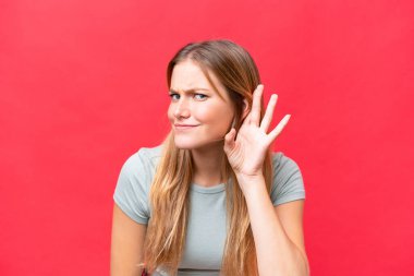 Young beautiful woman isolated on red background listening to something by putting hand on the ear