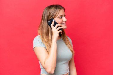 Kırmızı arka planda izole edilmiş genç ve güzel bir kadın cep telefonuyla birileriyle sohbet ediyor.
