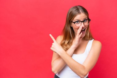 Kırmızı arka planda izole edilmiş genç ve güzel bir kadın bir ürün sunmak ve bir şeyler fısıldamak için kenarı işaret ediyor.