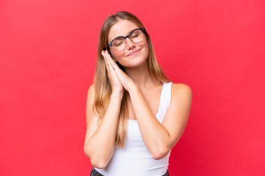 Genç, güzel bir kadın, kırmızı arka planda izole edilmiş bir şekilde uyku jesti yapıyor.