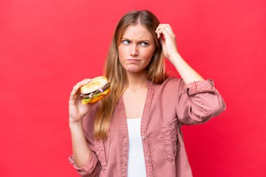 Kafkasyalı genç bir kadın elinde kırmızı arka planda izole edilmiş bir hamburger tutuyor. Şüpheleri var ve kafası karışmış bir yüz ifadesi var.