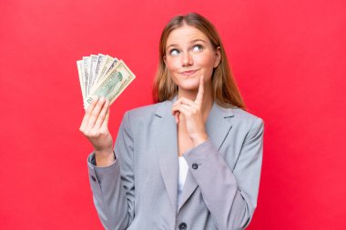 Young business caucasian woman holding money isolated on red background thinking an idea while looking up