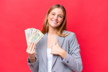 Young business caucasian woman holding money isolated on red background giving a thumbs up gesture