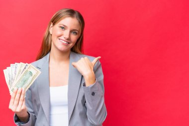 Young business caucasian woman holding money isolated on red background pointing to the side to present a product