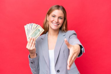 Kafkasyalı genç bir kadın kırmızı arka planda izole edilmiş parayı tutuyor ve iyi bir anlaşma için el sıkışıyor.
