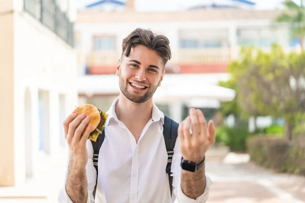 stock image Young handsome man holding a burger at outdoors inviting to come with hand. Happy that you came