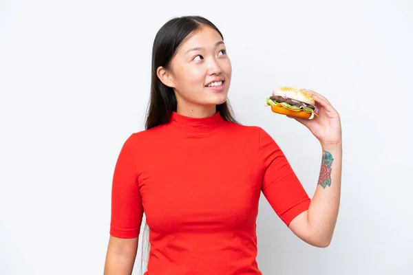stock image Young Asian woman holding a burger isolated on white background looking up while smiling