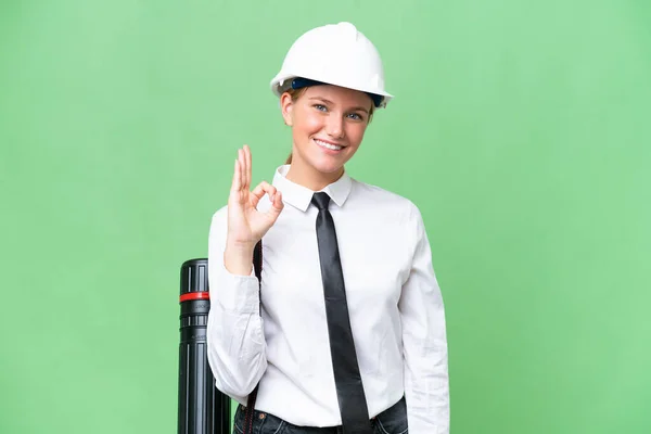 若い建築家Caucasian女性とヘルメットと保持設計図上の隔離された背景ショーOkサインとともに指 — ストック写真