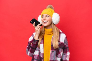 Kırmızı arka planda izole edilmiş kış maskesi takan genç ve güzel bir kadın cep telefonuyla sohbet ediyor.