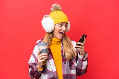 Kırmızı arka planda izole edilmiş kışlık kuklalar takan genç ve güzel bir kadın. Elinde kahve ve cep telefonu var.