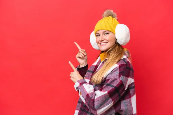 冬のマフを身に着けている若い美しい女性は 側に指を指して 製品を提示赤い背景に隔離 — ストック写真