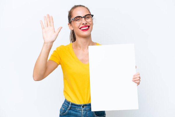 Young caucasian woman isolated on white background holding an empty placard and saluting
