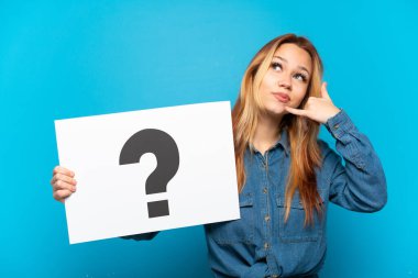 İzole edilmiş mavi arka planı olan genç bir kız. Elinde üzerinde soru işareti olan bir levha tutuyor ve telefon hareketi yapıyor.